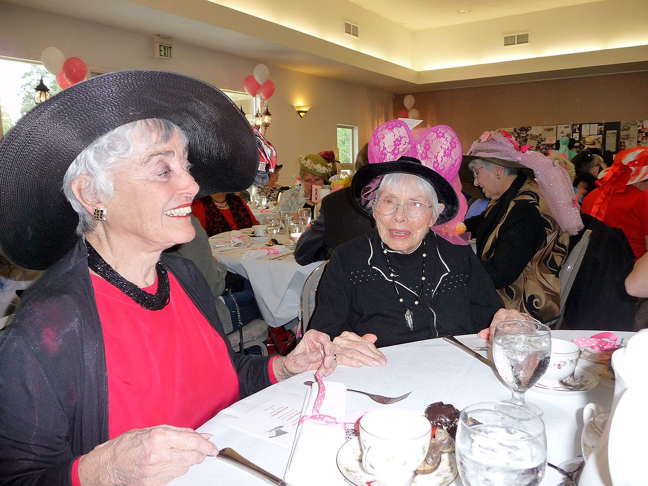 Patricia Morrison Coate/Olympic Peninsula News Group Thelma Bullock, left, is one of eight organizers of the Mad Hatters Tea Party. Seated next to her is 87-year-old Betty Newlon sporting her Betty’s Boobs hat and the message of “Fishing for a Cure.”