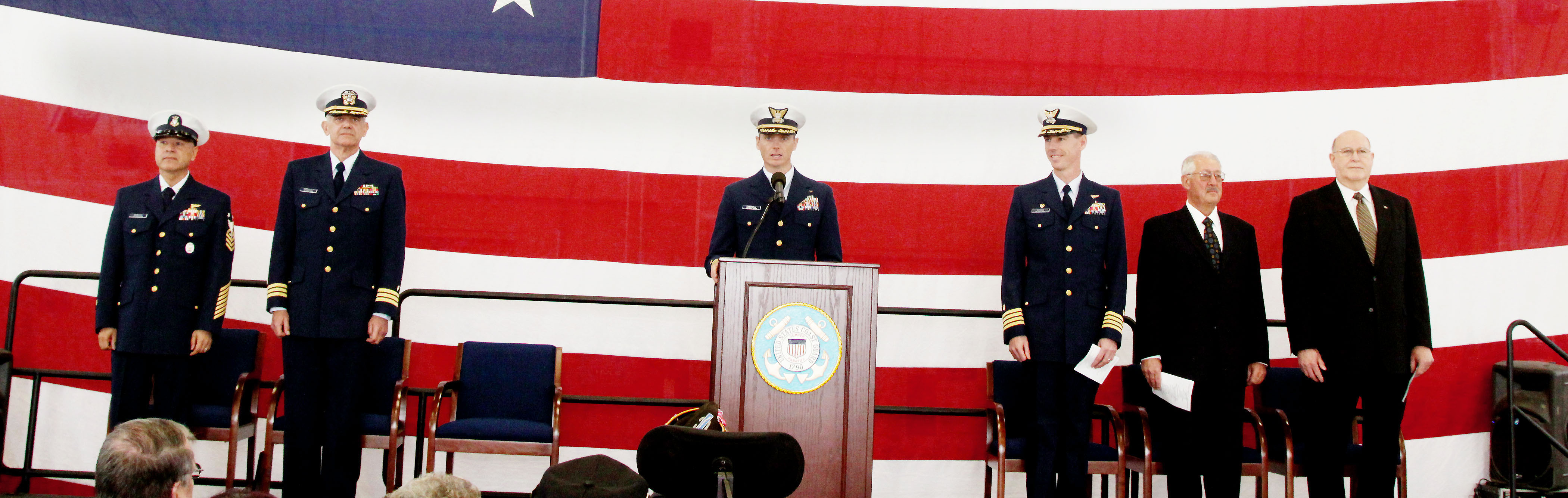 Ceremonies for the region's official Veterans Day ceremony at a hangar at Coast Guard Air Station/Sector Field Office in Port Angeles begin in front of a giant U.S. flag with station executive officer Cmdr. Michael Campbell at the podium. From left are Command Master Chief Petty Officer L.P. Moroles