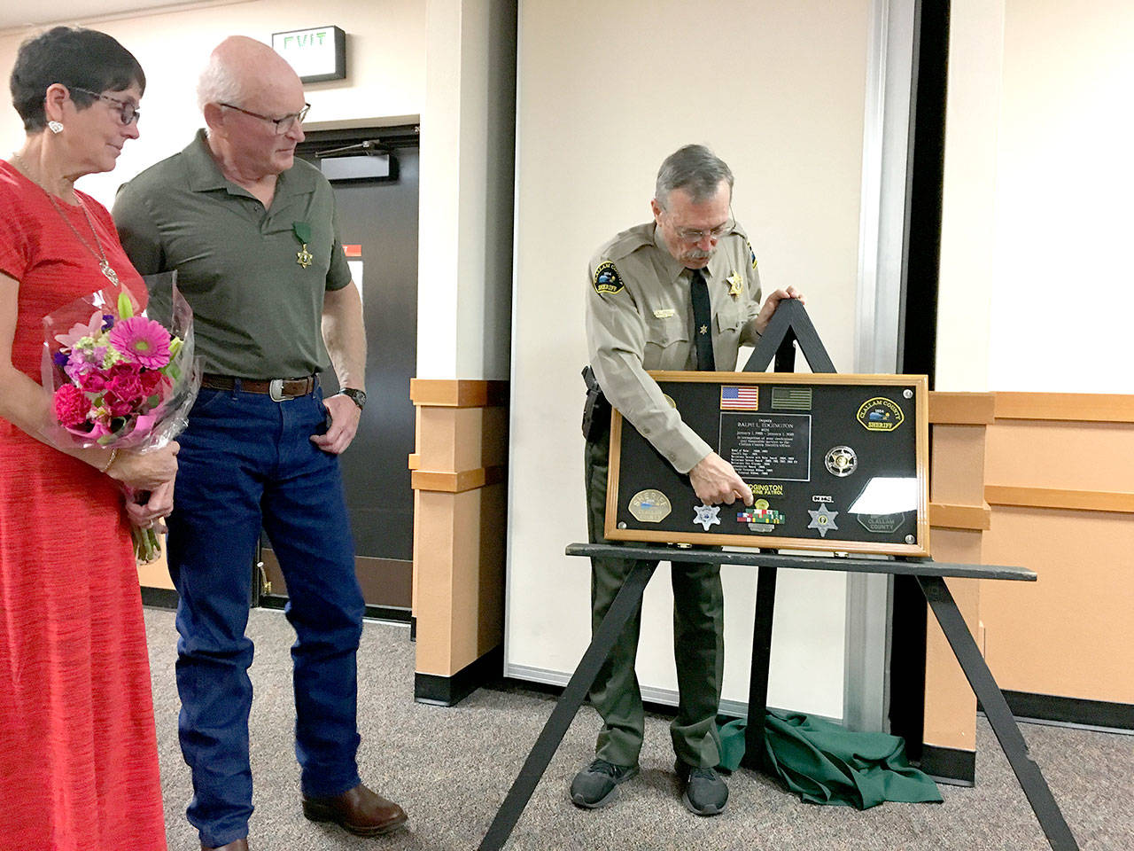 Retired Clallam County Sheriff’s Deputy Ralph Edgington and his wife, Karan, look on as Sheriff Bill Benedict displays a shadowbox depicting Edgington’s service medals. Edgington was recognized at the county commissioners’ meeting Tuesday for his 30 years of service to Clallam County. (Rob Ollikainen/Peninsula Daily News)