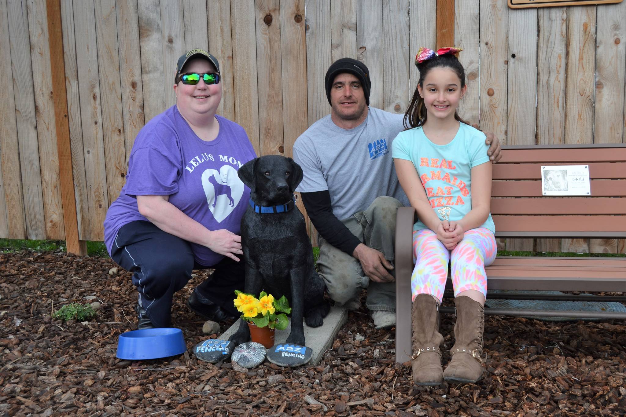 Jen Frame, co-moderator of the Sequim community page on Facebook, Kevin Cassidy and his daughter Alabama sit by the newest Stolli the dog statue in front of A2Z Fencing earlier this week. Cassidy recently installed the statue after community members raised funds to bring in a new statue after it was broken and stolen. Stolli was the Cassidys’ family dog who was popular among locals. (Matthew Nash/Olympic Peninsula New Group)