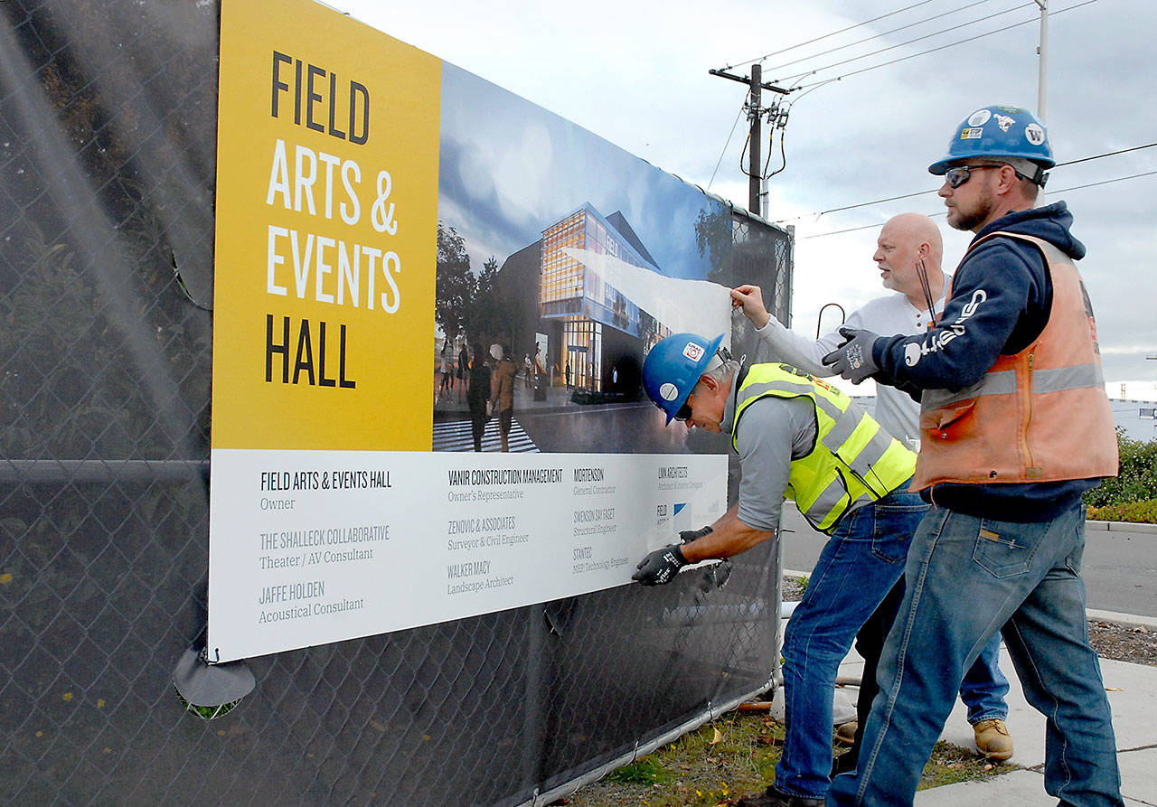 Chris Fidler, executive director of the Field Arts and Events Hall, center, gets assistance from Don Frostad, left, and Clayton Frohman of general contractor M.A. Mortenson Company as they peel back the protective cover from a project sign at the future site of the performing arts center at Front and Oak streets on the Port Angeles waterfront. (Keith Thorpe/Peninsula Daily News file)