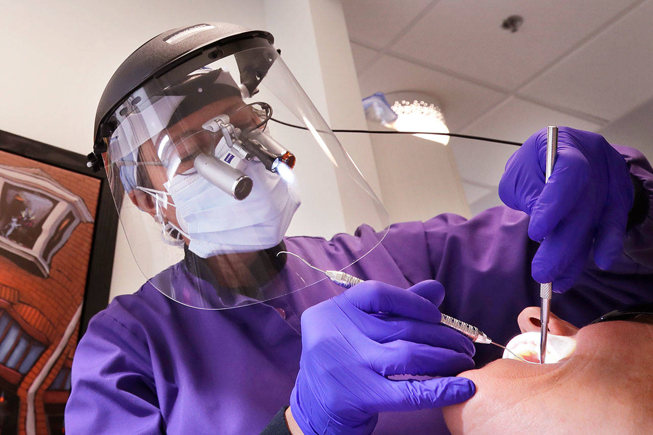 In this May 27, 2020, photo, dentist Dr. Kathleen Saturay wears additional protective equipment, including a face shield and disposable mask over a respirator mask, as she exams a patient in Seattle. Dental practices were allowed to open earlier this month following closure because of the coronavirus outbreak, as long as offices have enough personal protective equipment (PPE) for their staffers, put in place social-distancing policies and check patients for symptoms of COVID-19. (Elaine Thompson/Associated Press)