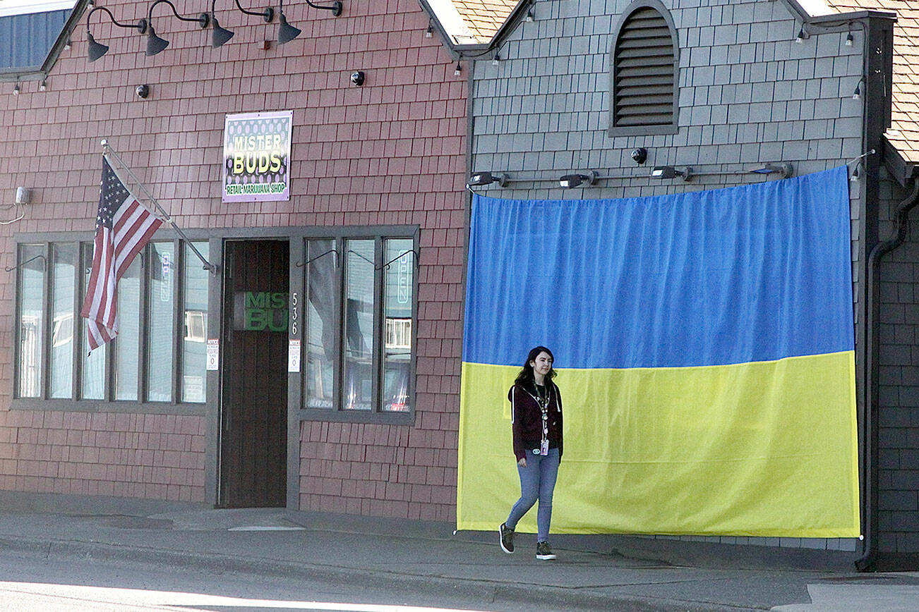 Shawna Small walks on Marine Drive in Port Angeles past a large Ukrainian flag on the outside of Mister Buds, which owner Malik Atwater put up recently along with the U.S. flag he has flown for many years. The flag was hand-sewn by Vivian Wai, Atwater’s wife, after the two discussed how to support the Ukrainians as they fight against the invasion of the country launched by Russia on Feb. 24. “We felt helpless to do anything to help,” Atwater said, adding that it was Wai who came up with the idea. (Dave Logan/for Peninsula Daily News)