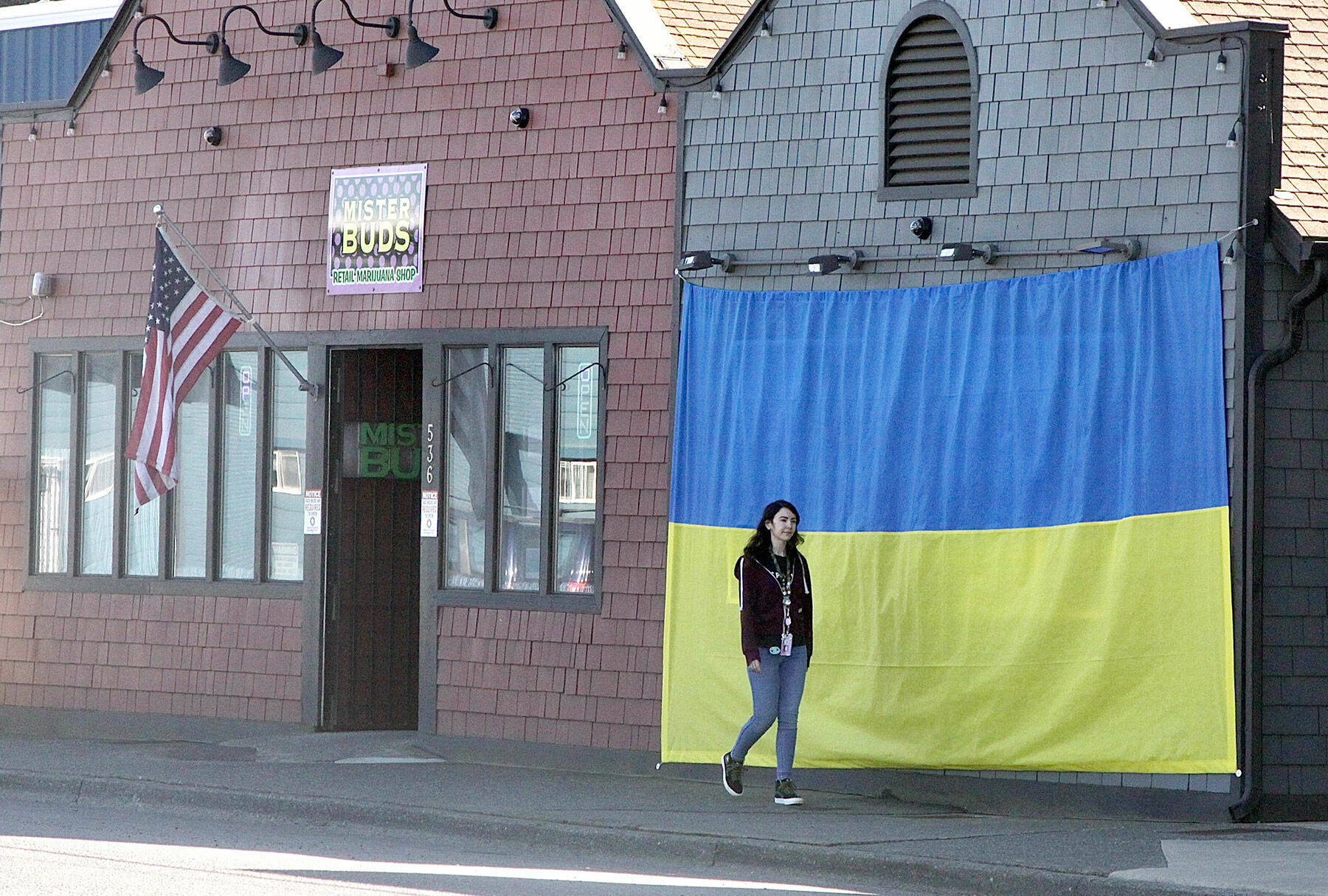 Shawna Small walks on Marine Drive in Port Angeles past a large Ukrainian flag on the outside of Mister Buds, which owner Malik Atwater put up recently along with the U.S. flag he has flown for many years. The flag was hand-sewn by Vivian Wai, Atwater’s wife, after the two discussed how to support the Ukrainians as they fight against the invasion of the country launched by Russia on Feb. 24. “We felt helpless to do anything to help,” Atwater said, adding that it was Wai who came up with the idea. (Dave Logan/for Peninsula Daily News)