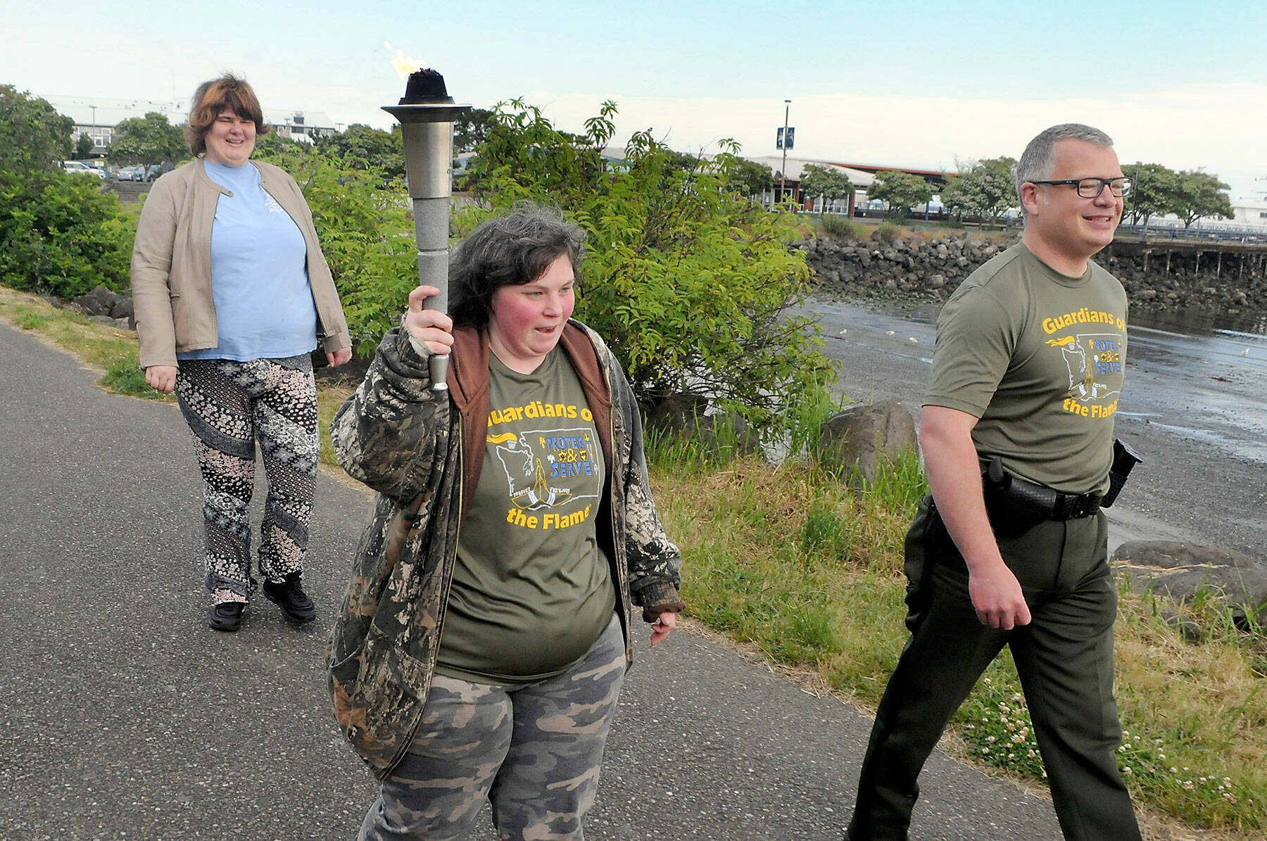 Special Olympian Deedra Hunter of Port Angeles takes a turn carrying the torch accompanied by fellow Olympian Bonny Ann Cates, left, and Chief Criminal Deputy Brian King of the Clallam County Sheriff’s Office during Wednesday’s Law Enforcement Torch Run along a section of the Waterfront Trail in Port Angeles. The relay, which included representatives from the sheriff’s offices in Clallam and Jefferson counties, Port Angeles and Sequim police, tribal police from Lower Elwha Klallam and Jamestown S’Klallam, State Patrol, Quilcene firefighters, U.S. Border Patrol, state parks personnel and Olympic National Park rangers, as well as several Special Olympians, followed a route from the west side of Port Angeles at 7 a.m. to the Hood Canal Bridge to about 5 p.m. in support of Special Olympics, which provides sports training and athletic competition for individuals with disabilities. (Keith Thorpe/Peninsula Daily News)