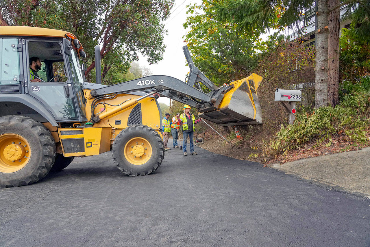 A road crew from the Port Townsend Public Works Department — Rafe Thornton, bucket driver, Tracy Benson, with hard hat, Chris MacDonald and Lane Dotson in the background — finishes repaving a section of Walnut Street on Thursday. The section of street was degraded for months with barriers set up to direct traffic around the worst part, essentially making it a one-lane roadway. The street was slated to be open for traffic later in the afternoon. (Steve Mullensky/for Peninsula Daily News)