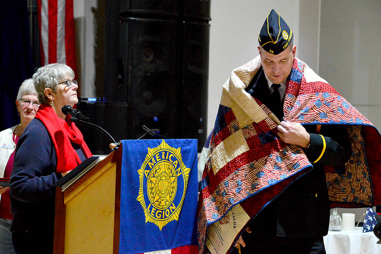 Kathryn Bates of North Olympic Peninsula Quilts of Valor, at the podium, helps present a quilt to U.S. Marine Corps veteran Wyatt Ranson on Friday. Ranson was one of 11 local veterans who received the honor on Friday at the American Legion Hall in Port Townsend. The Quilts of Valor volunteers make and present quilts to veterans of all branches of service in both Clallam and Jefferson counties. To request a quilt for a veteran, visit www.qovf.org or email 1katheybates@gmail.com. (Diane Urbani de la Paz/for Peninsula Daily News)