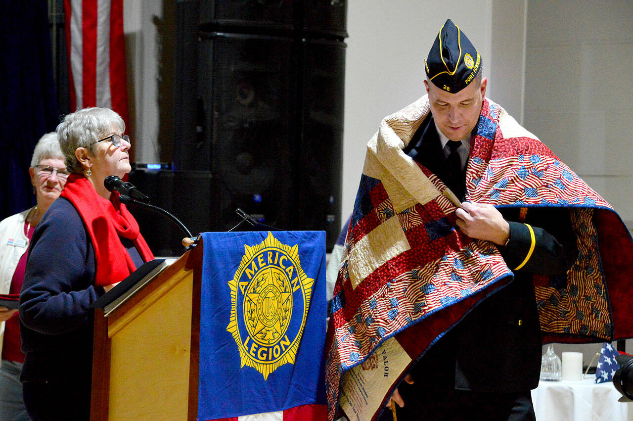 Kathryn Bates of North Olympic Peninsula Quilts of Valor, at the podium, helps present a quilt to U.S. Marine Corps veteran Wyatt Ranson on Friday. Ranson was one of 11 local veterans who received the honor on Friday at the American Legion Hall in Port Townsend. The Quilts of Valor volunteers make and present quilts to veterans of all branches of service in both Clallam and Jefferson counties. To request a quilt for a veteran, visit www.qovf.org or email 1katheybates@gmail.com. (Diane Urbani de la Paz/for Peninsula Daily News)