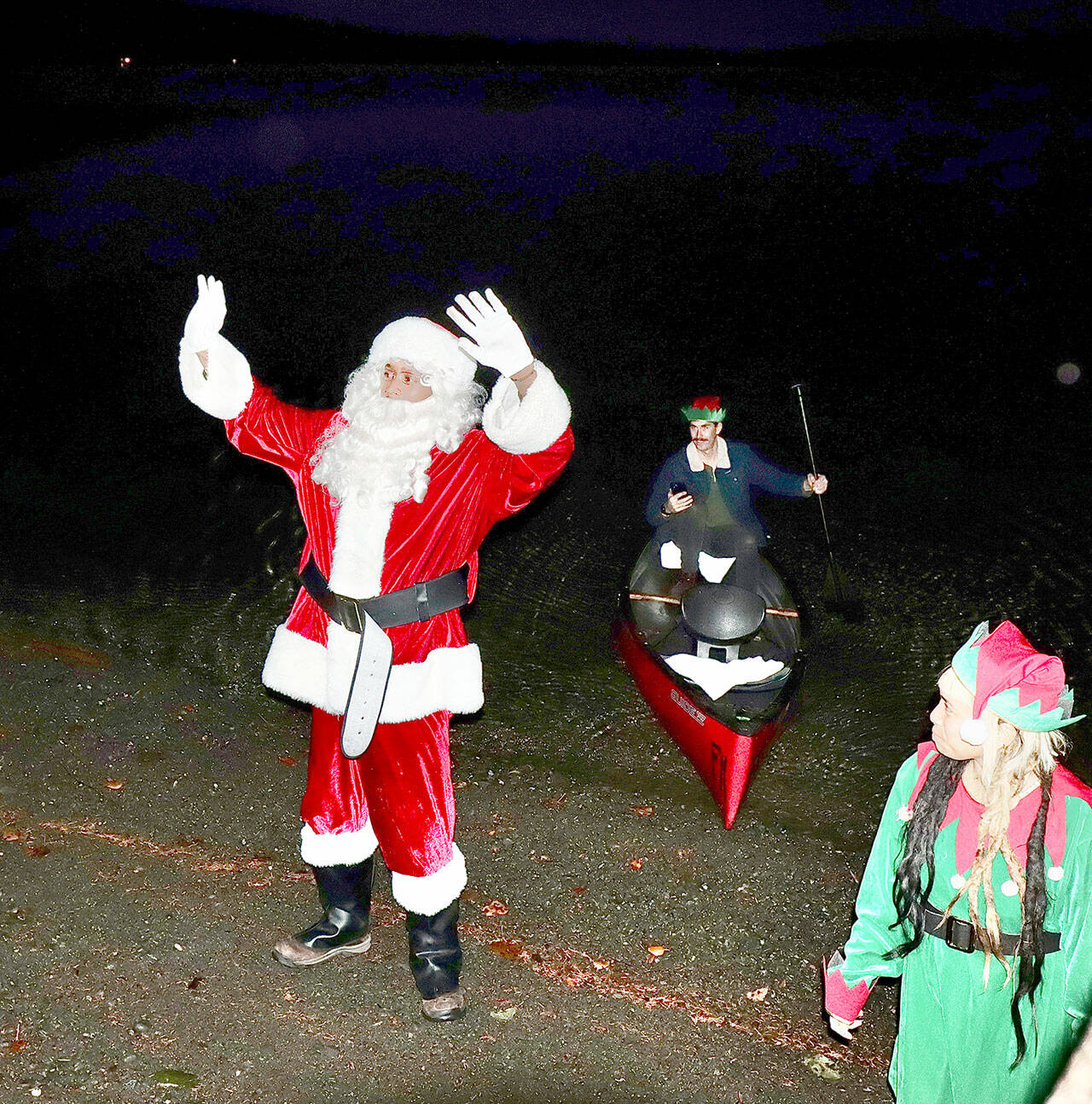 Santa, played by Beau Gibson, greets children and their parents on the shores of Lake Crescent after his arrival by canoe to the Lake Crescent Lodge on Saturday. In the canoe is his paddler Cody Fisher and the elf on the shore is Vera Tambunan. (Dave Logan/for Peninsula Daily News)