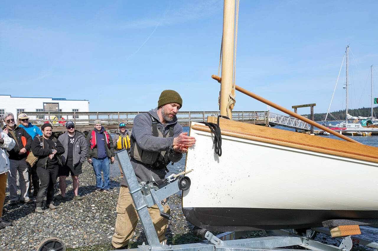 Students watch as Northwest School of Wooden Boatbuilding instructor Korey Ruben splashes sea water on the hull of a recently completed 14-foot Cat boat built at the school and launched on Thursday. A Cat boat is a style that originated in the northeast and sailed around Martha’s Vineyard often as a party boat because it is wide enough to accommodate several people. (Steve Mullensky/for Peninsula Daily News)