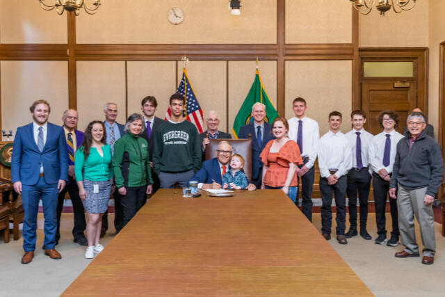 Gov. Jay Inslee, center, signs Substitute Senate Bill No. 5687 on April 25 — legislation called the Charles Cate II Act that bolsters the creation and support of postsecondary wrestling grant programs. Joining Inslee are Cate's son Charley and wife Renee. Behind Inslee is primary bill sponsor Sen. Kevin Van De Wege and Sequim wrestling advocate Randall Tomaras, along with wresters and wrestling advocates. At far right is SHS wrestling coach Steve Chinn. (Randy Tomaras/Gov. Jay Inslee's office)