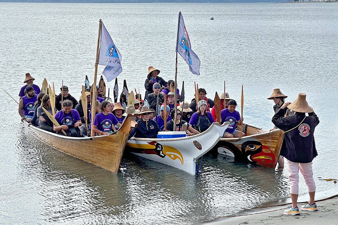 Celeste Dybeck, elder with the Jamestown S’Klallam Tribe in Sequim, invites the pullers from the Quileute Tribe in La Push to come ashore at Fort Worden in Port Townsend and rest and eat before continuing the Paddle to Muckleshoot for the annual Tribal Canoe Journey. The tribal members are wearing purple, the color of the Missing and Murdered Indigenous Women and Children movement, to honor the memory of tribal member Virginia Castaneda, 19, who was murdered in a domestic violence dispute about six years ago. An empty seat was left for her in one of the canoes. The stop was the final one on the North Olympic Peninsula before canoes are due to reach their destination on Sunday. (Steve Mullensky/for Peninsula Daily News)