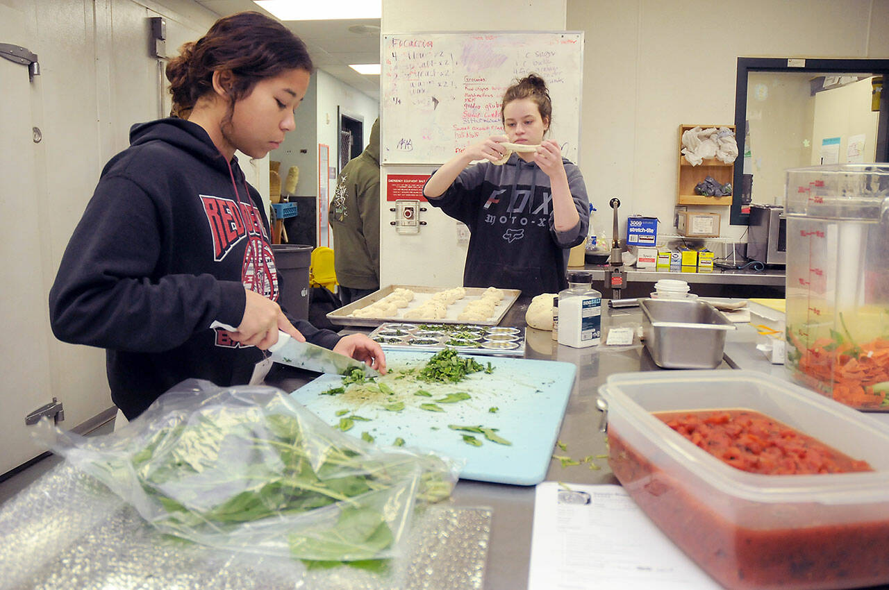 Salish Sea Ecotourism and Hospitality student Julia Livingston, 17, left, dices basil while Trinity Williams, 18, creates garlic bread twists at the Port Angeles School District’s commercial kitchen classroom. (KEITH THORPE/PENINSULA DAILY NEWS)<address><em></em></address>