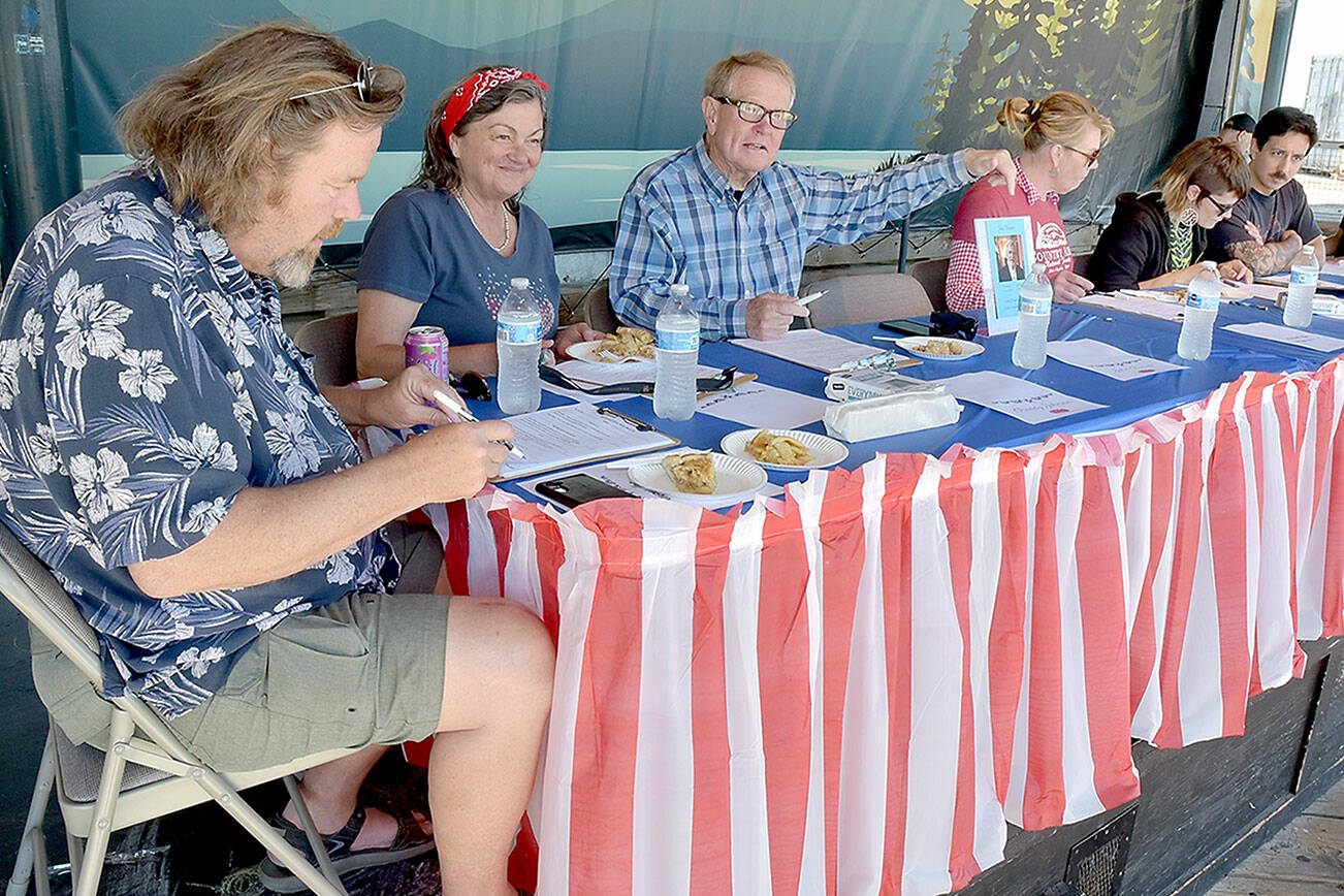 Pie contest judges, from left, Jeff Tocher, Laurel Hargis and Clallam County Commissioner Randy Johnson, along with event sponsor Country Aire Natural Foods representatives Kristina Fallon, Katie Meyers and Caeron Alarcon, make their choices during Thursday’s annual Independence Day apple pie contest at Port Angeles City Pier. A total of 26 pies were entered into the contest with first-place honors going to Xaven McCarty of Seattle, Elisabeth Pennell of Mukilteo in second and Selena Reach of Port Angeles taking third. The contest was dedicated the late legacy judge and former Peninsula Daily News publisher John Brewer, represented by a photo in front of an empty chair. (Keith Thorpe/Peninsula Daily News)