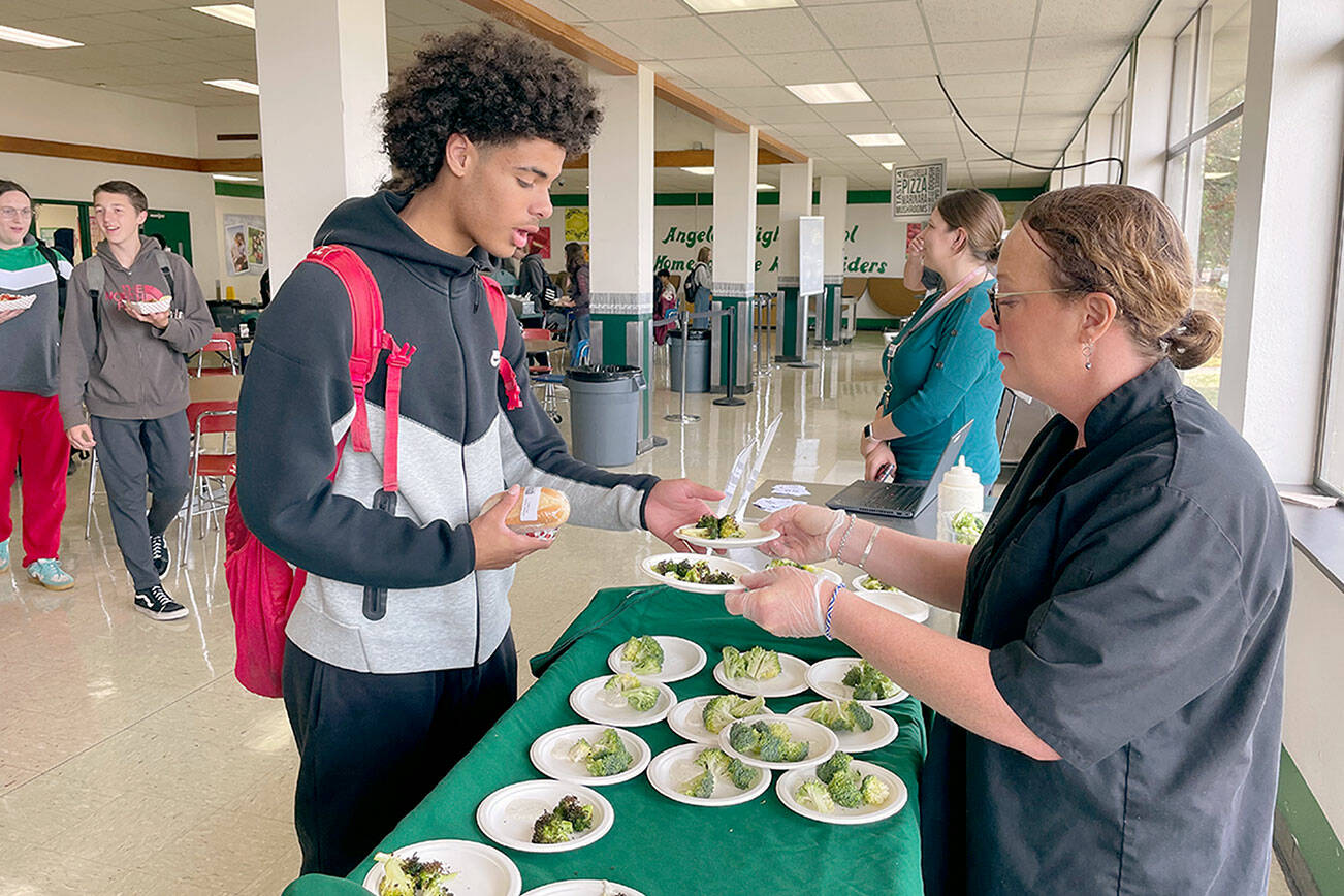 Port Angeles High School junior Tucker Swain, left, tries out a sample of roasted broccoli with ranch dressing dipping sauce prepared by Stacey Larsen, the district’s WSU Clallam Extension Farm to School consultant at the school’s cafeteria on Friday. Including locally grown produce like the Chi’s Farm broccoli into meals, increasing the amount of whole grains in foods and reducing salt and added sugar are part of the school district’s efforts to create healthier options and meet updated USDA nutrition standards. A new app provides students and parents a way to view menus and the nutritional content, calories and allergens in meal options. (Paula Hunt/Peninsula Daily News)