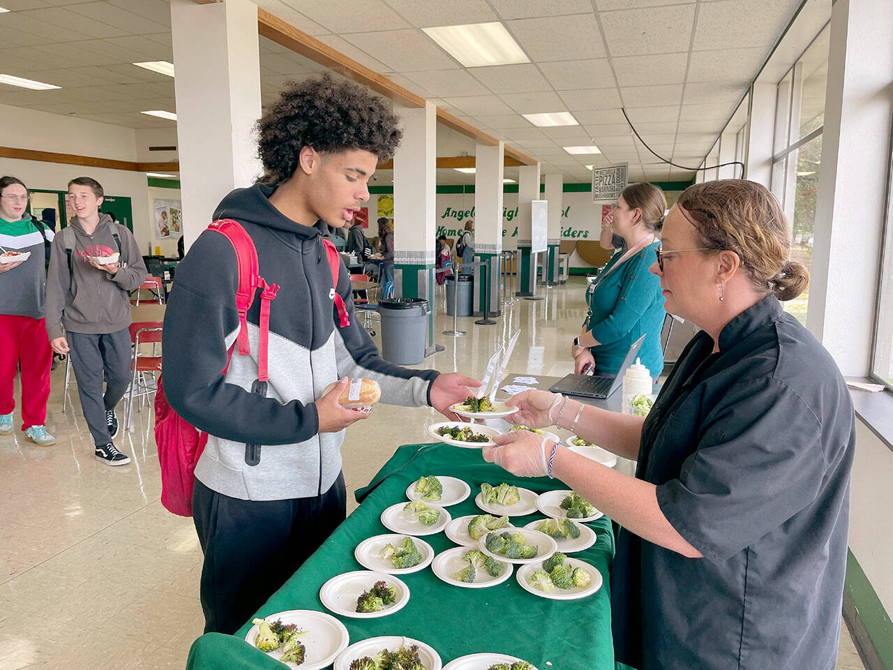 Port Angeles High School junior Tucker Swain, left, tries out a sample of roasted broccoli with ranch dressing dipping sauce prepared by Stacey Larsen, the district’s WSU Clallam Extension Farm to School consultant at the school’s cafeteria on Friday. Including locally grown produce like the Chi’s Farm broccoli into meals, increasing the amount of whole grains in foods and reducing salt and added sugar are part of the school district’s efforts to create healthier options and meet updated USDA nutrition standards. A new app provides students and parents a way to view menus and the nutritional content, calories and allergens in meal options. (Paula Hunt/Peninsula Daily News)