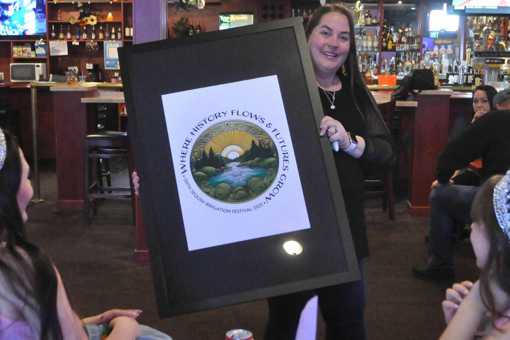 Michelle Rhodes, executive director of the Sequim Irrigation Festival, shows festival royalty the new logo and tagline “When History Flows and Futures Grow” for the 130th festival on Oct. 12 at the Oasis Bar and Grill. Sherry Scharschmidt designed the logo using artificial intelligence technology. (Matthew Nash/Olympic Peninsula News Group)