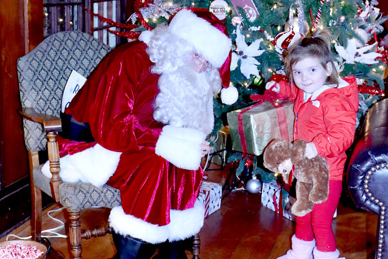 Santa, played by Alexis Smith, a Lake Crescent Lodge employee, wonders why Zoey Doud, 3, of Port Angeles thinks the big present under the tree is for her. Santa turned on the outdoor lights at the lodge on Saturday then sat down by the fireplace to visit with the many children who came to the festivities. Entertainment was provided by the holiday songs of the Grand Olympic Chorus. (Dave Logan/for Peninsula Daily News)