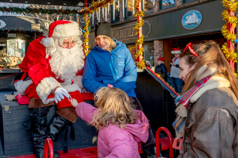 Sula Adams, 4, hands Santa a candy cane during Santa’s visit to Port Townsend. Santa made an early visit on Saturday to hear children’s Christmas wishes and to light the community tree at Haller Fountain. Sitting next to Santa is Sula’s grandma Christi and her mom Corrine is on the right. (Steve Mullensky/for Peninsula Daily News)