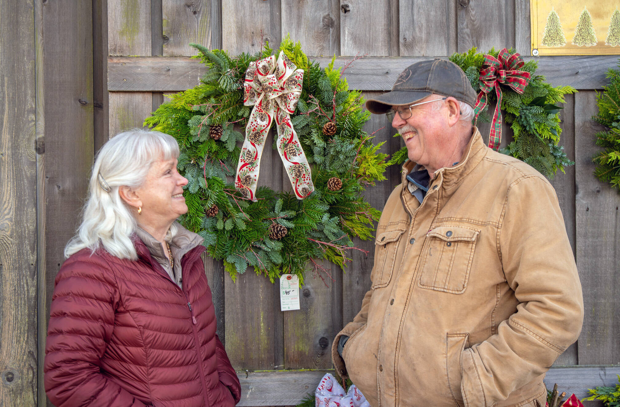 Outside the Christmas Barn at Lazy J Tree Farm, Ann and Steve Johnson smile at each other in front of wreaths Ann prepares with two other women for the Christmas season. (Emily Matthiessen/Olympic Peninsula News Group)