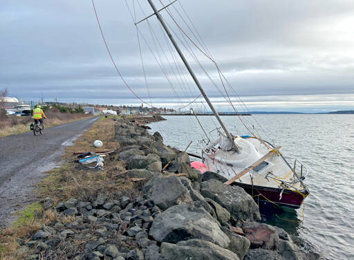 A cyclist rides by the 26-foot sloop that was dashed against the rocks along the Larry Scott Trail on Wednesday due to 30 mph winds from an atmospheric river storm buffeting the North Olympic Peninsula. A 29-year-old Port Townsend man, who was not identified, and his dog were rescued by a Coast Guard rescue swimmer from Coast Guard Air Station Port Angeles. (Steve Mullensky/for Peninsula Daily News)