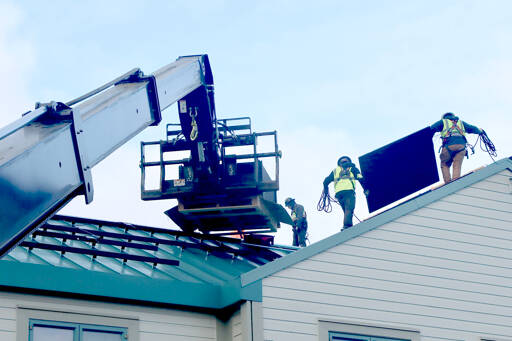 C.J. Conrad and Chris Orr of A&R Solar take solar panels from a lift on top of the Port Angeles Senior and Community Center on Peabody Street to be installed on the roof. The 117 panels are mostly made of silicone and will provide electrical power to the center. The crew members are each tied in with ropes to prevent any problems on the slippery slanted roof. The panels are 42 inches by 62 inches and weigh about 16 pounds. (Dave Logan/for Peninsula Daily News)