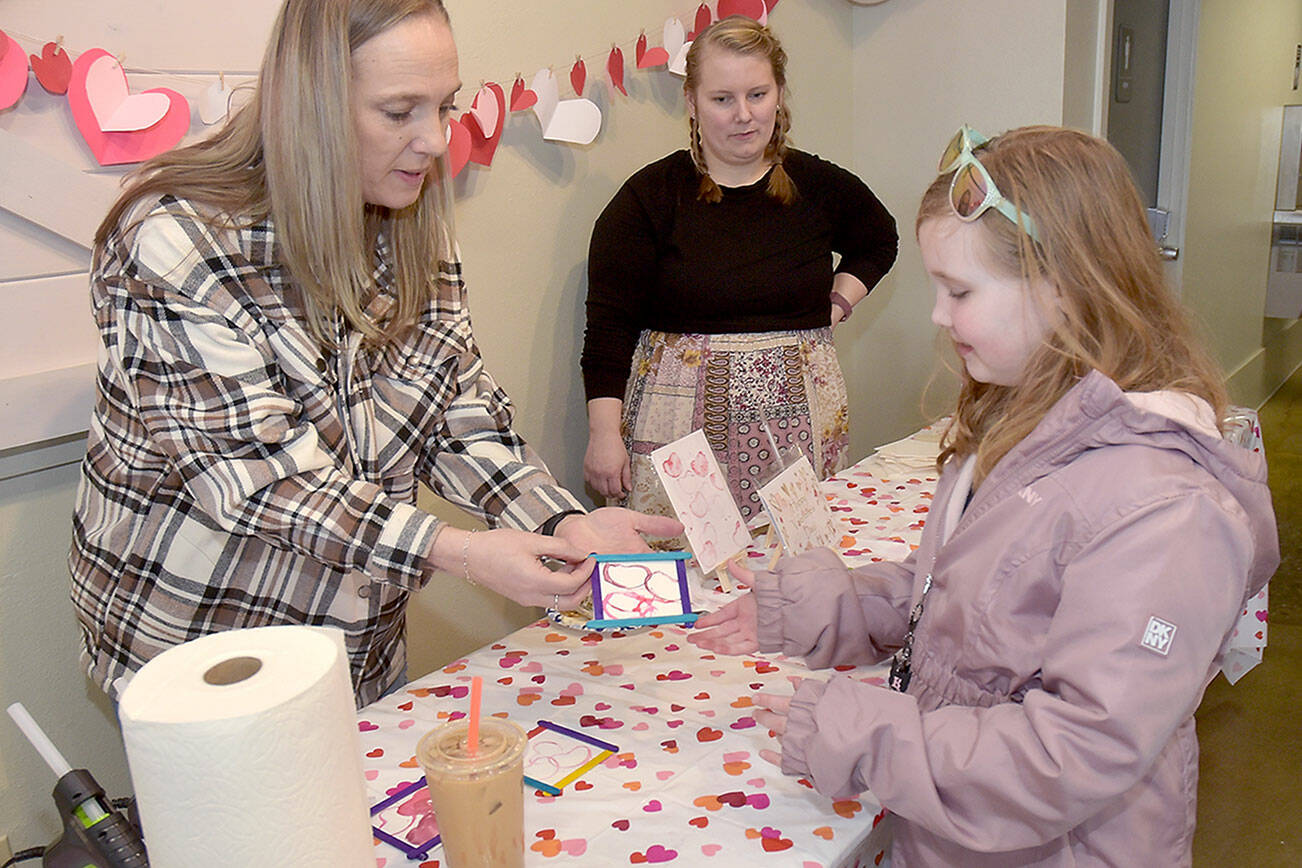 Madison Canterbury, 7, of Shelton looks at a Valentine’s gift she created with the assistance of volunteers Chandra McGuff, left, and Nicole Lemon at a craft table set up outside the Silver Lining Cafe at The Wharf on Saturday on the Port Angeles waterfront. Youngsters were given the opportunity to create gifts and Valentine’s cookies during the event. (Keith Thorpe/Peninsula Daily News)