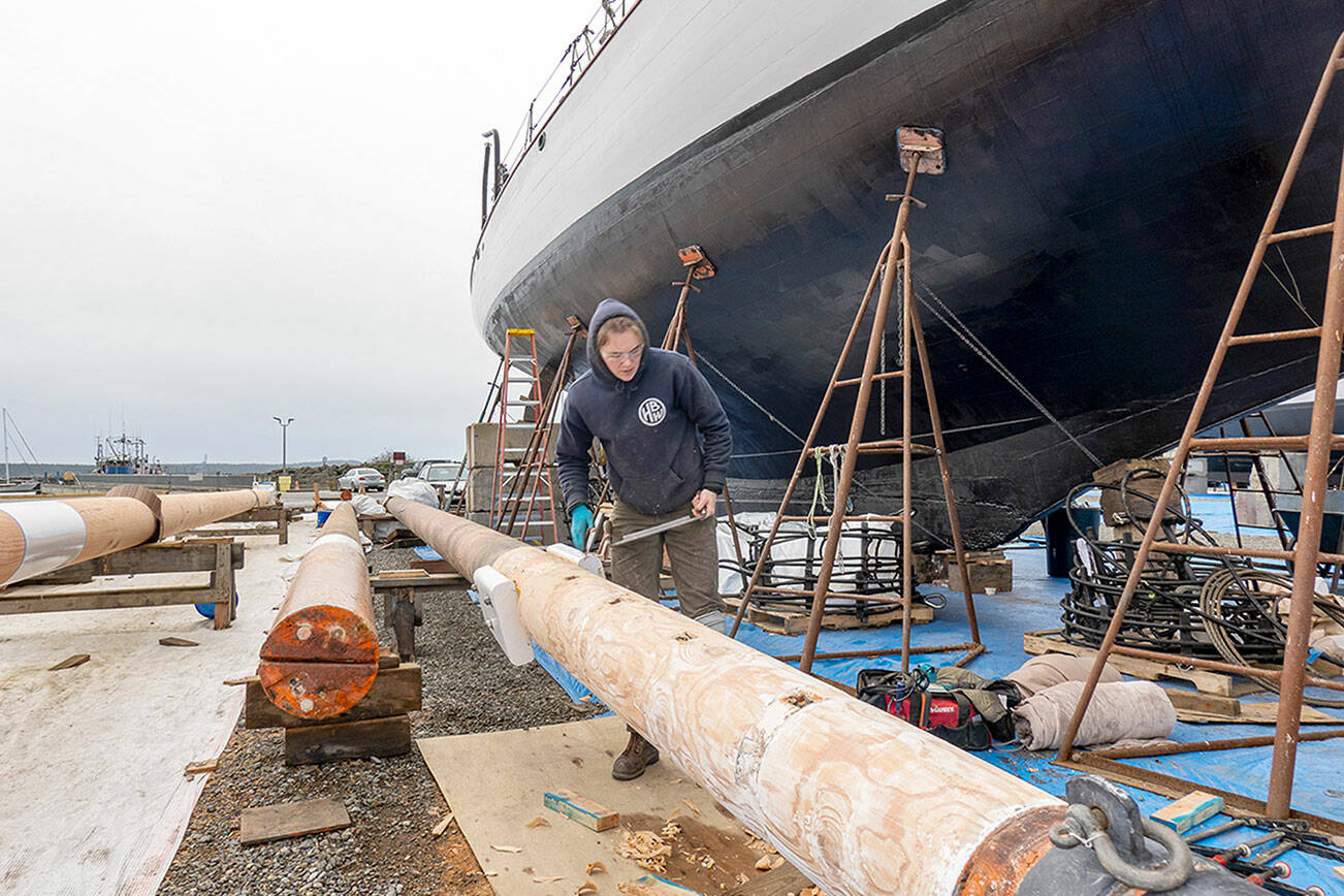 Zoe Hewitt, a shipwright at Haven Boatworks, uses a draw knife to shape the foremast on the 112-year-old historic schooner Adventuress, background, that is resting on the hard at the Port Townsend Marina. A new main mast, left, and the fore mast will be stepped next week, weather permitting. (Steve Mullensky/for Peninsula Daily News)